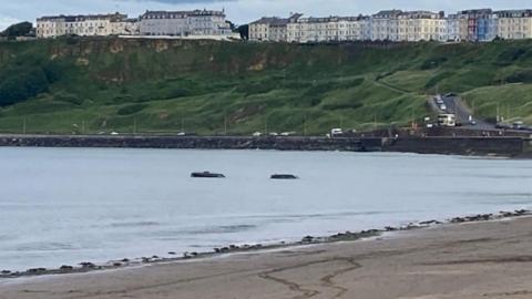 Two cars stranded in the sea in Scarborough. There are cliffs in the background with a series of coloured houses on top.