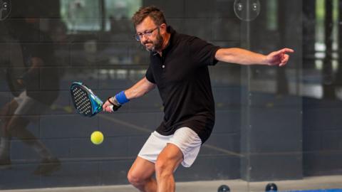 A man with a stern look about to hit a forehand with a padel racket, at an indoor centre. He has black hair, a black and grey beard, glasses and a necklace. He is wearing a black top and white shorts.