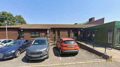 A google street view image of a single storey office building with and orange car, silver car and blue car parked outside.