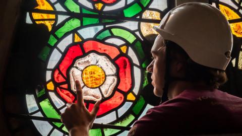 Apprentice glazier Theo Dives, from York Glaziers Trust, wears a white hard hat. He points to a small section of the Rose Window's stained glass in the shape of a  red, white and yellow rose