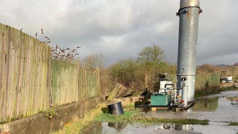 An outdoor area showing a partially collapsed fence standing next to a long metal chimney. A tarmacked area with sporadic patches of grass can be seen, with puddles across the surface.