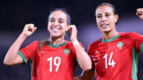 Sakina Ouzraoui and Aziza Rabbah wear red Morocco shirts with green trim, smile and raise their hands in the air in celebration