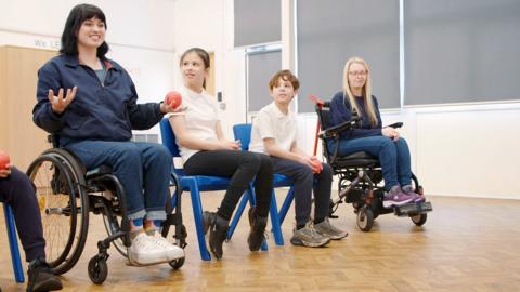 Blue Peter presenter, children and school staff in a gym hall learning how to play boccia.