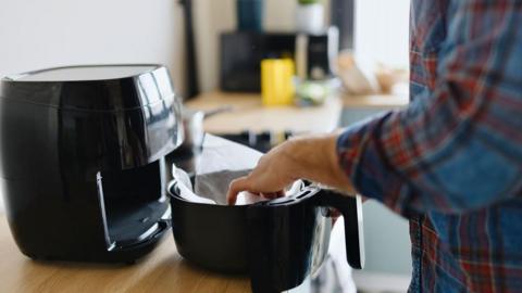 A black air fryer on a wooden kitchen work top. Part of it has been pulled out with someone placing some clear paper into it. The person is wearing a blue and red tartan shirt