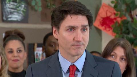 Justin Trudeau wells up during one of his last press conferences as leader of Canada. He is wearing a steel blue suit, red tie and blue shirt. Four people are visible standing behind him.