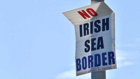 A sign on a lamppost in red white and blue lettering says No Irish Sea Border with unionists against NI Protocol on smaller writing at the bottom