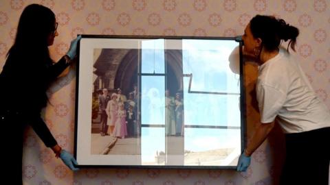 Two women wearing gloves lift a framed photograph of a family portrait on to a gallery wall, which is wallpapered. 