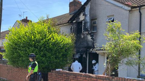 A terraced two-storey house shows fire damage on the groundfloor, first floor outer wall, window, part of the roof. Two people in a forensic outfit are at the front door whole a police woman is guarding the property from outside a waist-high brick wall.