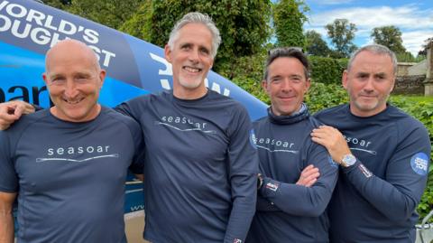 Four teammates wearing navy blue tops stand in front of a blue boat. They are smiling and have their arms around each other. 