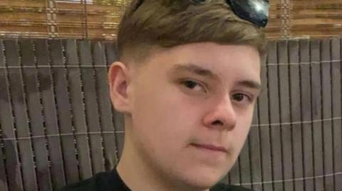 A teenage boy with dark hair and sunglasses on his head. He is starting into the camera. A fence is behind him.