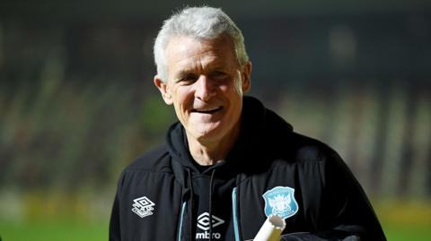 Mark Hughes smiles, wearing a black Carlisle coat and tracksuit