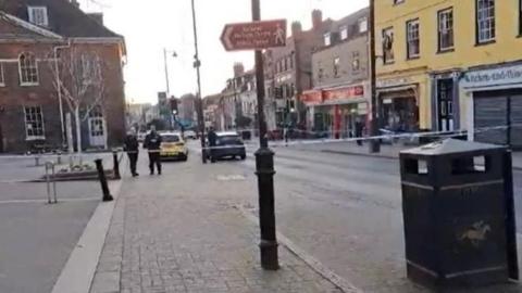 High Street in Newmarket, police officers standing close to a police car. There is police tape across the road, there are buildings on either side of the road 