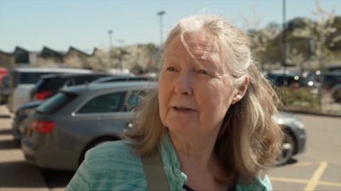 A woman with shoulder length grey hair looking at the camera wearing a turquoise jacket with a pale grey bag strap visible. She is stood in a car park with a silver car directly behind.