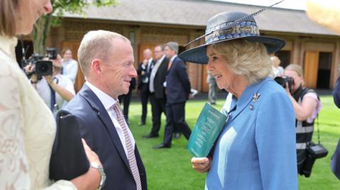 Several people are gathered outdoors on a lawn in front of a brick building during a formal event. In the foreground, two people are engaged in conversation. One person is wearing a light blue jacket and a wide-brimmed dark hat decorated with ribbon and a feather, and is holding a turquoise event programme. The other person is wearing a dark suit with a patterned tie. In the background, additional attendees in formal attire are standing and talking, while photographers and a camera operator capture the event. The scene is brightly lit by sunlight, with green grass and trees adding to the setting. Visible text on the programme is too small to read clearly.