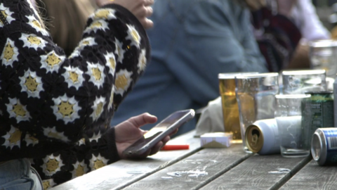 A table suggestive of an outdoor alcohol-vending recreational establishment with several pint glasses, one full,  the rest empty and some cans, some on their side, and a cigarettes packet. A person is wearing a knitted top on the left raising their right hand.