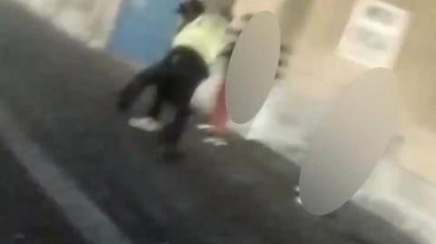 Image of a police officer in yellow arresting a teen people are blurred around the pair