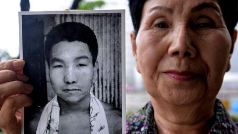 Hideko Hakamata wearing a floral shirt holds up a black-and-white photograph of her brother in her right hand, Iwao Hakamata, as a young man