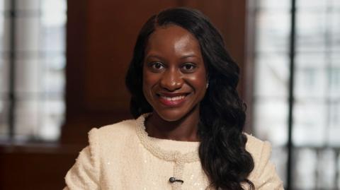 Afua Kyei wears a white suit jacket with a beaded trim. She has long dark hair and is smiling.