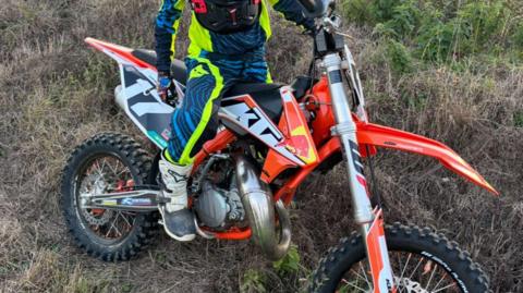 An orange and silver KTM motocross style bike. It's on a marsh land. The body of a boy can be seen posing with it. He's wearing blue and neon green clothing.