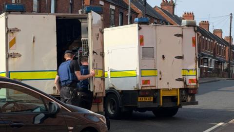 Two large white police vans, with two officers facing away from the camera wearing blue vests and berets. 