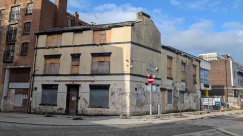 The Tithebarn Pub is an old cream-coloured two-storey building. Its windows are boarded up and the paint work is neglected and cracking. It is surrounded by temporary metal fencing.
