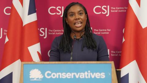 Kemi Badenoch gives a speech in front of a podium reading: "Conservatives". She is standing in front of a red backdrop reading: "CSJ The Centre for Social Justice" and two union jacks.