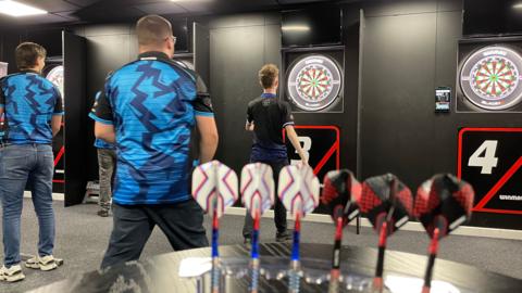 Four men stand in front of dart boards on a black wall at a newly built academy. Two of the men are standing right in front of the dart boards, while the other two watch from further back. There are several darts on a table in the foreground.