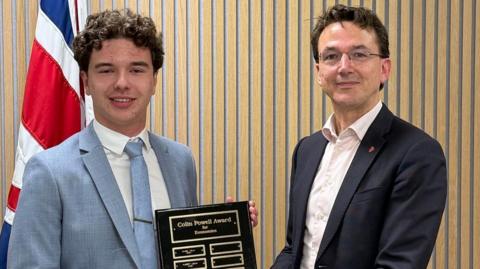 Two people standing indoors against a light wooden panelled wall. One person is holding a dark plaque with gold lettering that reads “Colin Powell Award” and a list of engraved name plates beneath. A second person beside them is helping to present the award.