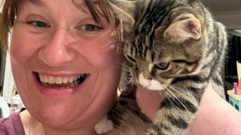 Marie Fox holding up Daisy the cat. Marie is smiling at the camera