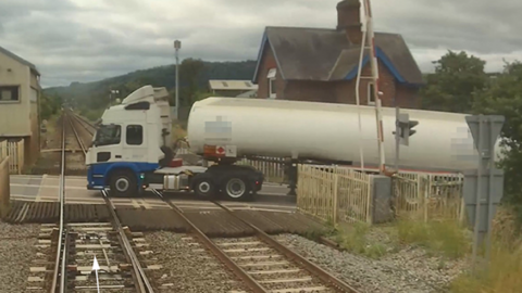 A white and blue tanker truck is pictured halfway across a railway level crossing. The company details on the sides of the vehicle have been blurred