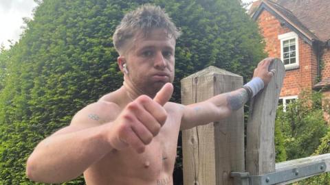 Tommy, a young man, has his top off and is doing a thumbs up at the camera with a tired look on his face. He is resting his other hand on a wooden gate by a house. He has several tattoos, including a cross on his sternum.