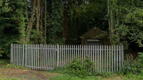 A single-storey brick building set back in woodland, with a metal security fence in the foreground 