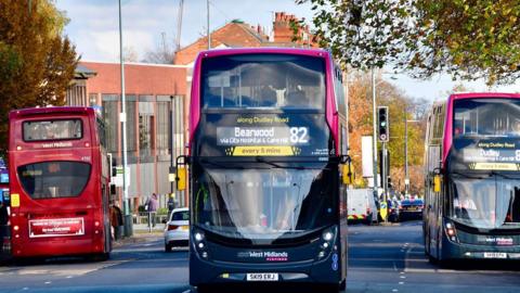 A three-lane road with three buses on it. A red bus is at a bus stop facing away from the camera. Another bus, with Dudley marked as its destination, is at a bus stop on the other side of the road facing the camera. Between them, a bus heading for Bearwood is driving towards the camera.