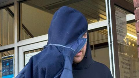 A close-up image of a man wearing a black hooded top with the hood of a navy coat pulled over his head and part of his face as he tried to evade the camera.