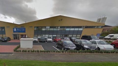 Yellow-painted industrial unit with mainly glass frontage and pointed roof. Name over the door says "Kettle Interiors".  There is a car park outside with most of the spaces full. 