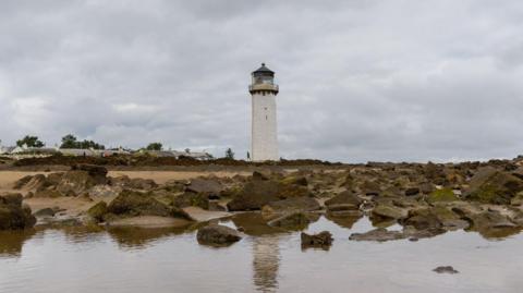 A white lighthouse is set against a grey sky. It sits on a sandy beach, but there are rocks in the foreground, and shallow water.