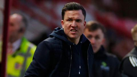 Exeter City boss Gary Caldwell, wearing a navy blue gilet over a navy blue hoodie, shouts instructions from his technical area