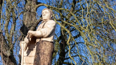 Image shows a tall wooden statue in front of a tree, on a bright sunny day. The statue is of a man, dressed in 1800s attire, standing with his arms crossed.