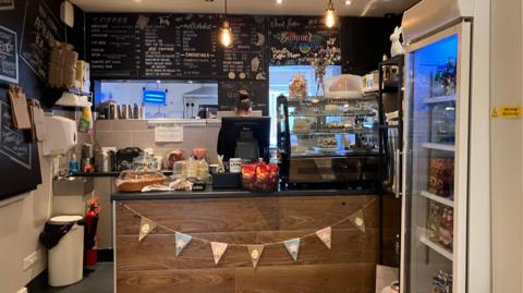 A photo of a cafe counter with loads of cakes on the counter there are also crisps on the counter. There are colourful flags hanging from the counter.
