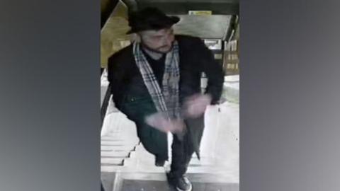 CCTV footage of a man of a man in an all black outfit - black shoes with a white sole, black trousers, a black long coat and black trilby-style hat. He is also wearing a beige check scarf and has a brown, trimmed beard. 