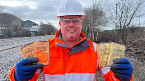 Rob Cochrane is standing by a railway line holding four video cassettes in his hands. The boxes are gold with black lettering. He is wearing an orange hi-vis jacket, a white safety helmet and blue gloves.