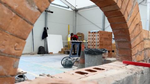 An archway of bricks inside a factory. Through the gap you can see a young man working at a bench with a pack of bricks by his side