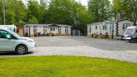 The Hut Lane traveller site showing two large one-storey homes in cream with white bow windows by a large gravel area with grass to the front. There are cars and mobile caravans to the wides of the photo
