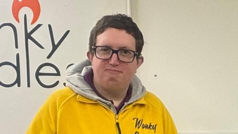 PJ Seaman holding one of his candles wearing a yellow Wonky Candles zip-up fleece