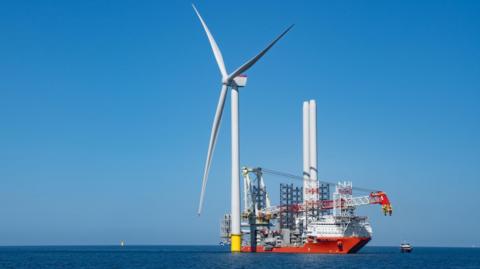 A wind turbine in the sea. Next to it, there is a large ship being used in the turbine construction process. The blue sea is flat and calm. The cloudless sky is blue.