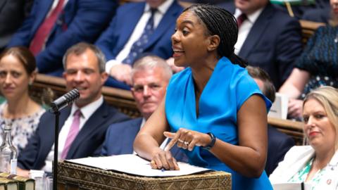 Kemi Badenoch in the House of Commons at the despatch box. She is wearing a blue dress and behind her are Conservative MPs.