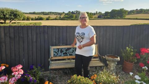 A woman with blonde hair in a ponytail and black glasses is smiling. She is standing in front of a wooden bench, which is in front of a fence - beyond which are an expanse of farmland fields and trees. In the garden in front of here are flowers and plants, as well as a bird bath