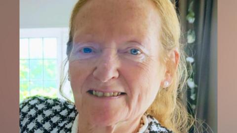 A woman in her late 60s, with blue eyes and light ginger hair tied back, smiles at the camera. She is inside a house with a green curtain and window behind her, and is wearing a black and white chequered jacket.