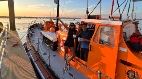 Jemma and her father stand on the deck of an orange lifeboat, which is docked in a harbour. In the background, open ocean can be seen, with the sun setting.