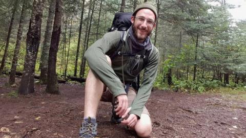Alex Pretti kneels in front of the camera wearing hiking gear in a green forest.
